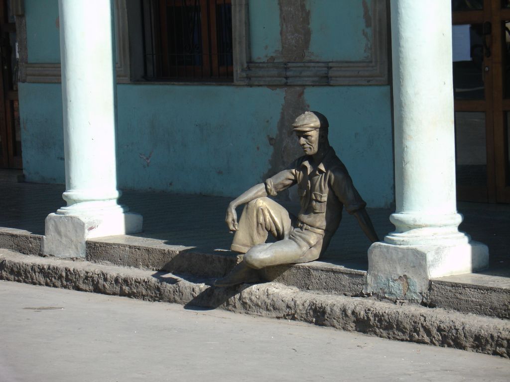 Dieser Mann ist eine Statue. Er sitzt vor einer Ladenzeile.