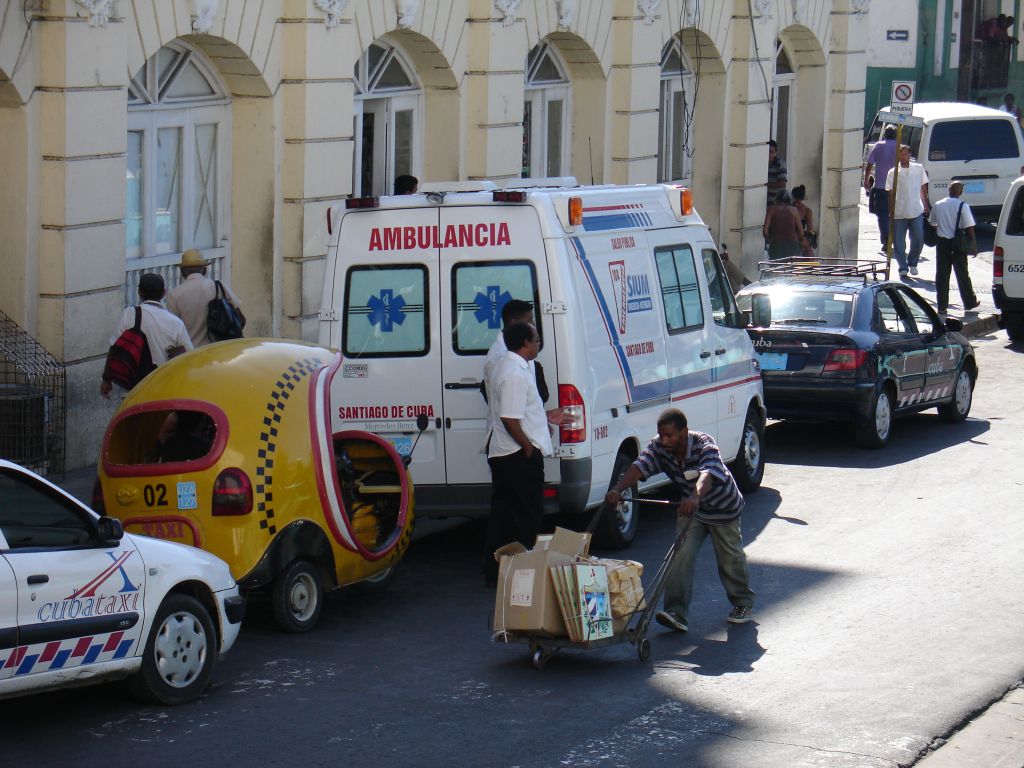 Ein Markthändler transportiert Bilder und andere Touristensouvenirs auf einem Handwagen.<br />Dahinter ein modernes Taxi, ein 'Cocotaxi' und ein Krankenwagen.