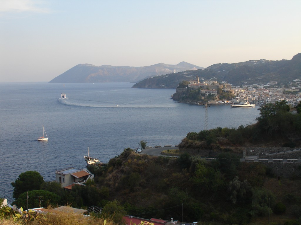 Der Ort Lipari hat 5000 Einwohner und ist der Hauptort der ganzen Inselgruppe. Die beiden Häfen des Ortes befinden sich beiderseits des Burghügels, der vor der Stadt im Meer liegt. Im Hintergrund sieht man die Insel Vulcano, von der die Vulkane weltweit ihren Namen haben.